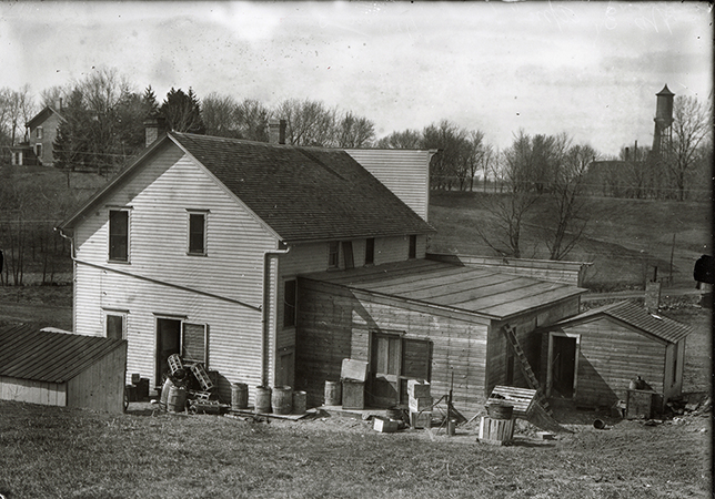 Photograph of the rear of the original Champlin store and livery located at the corner of Lincoln Way and Welch. In the background Marston Cottage can be seen to the left (north-west) while to the right (north) the Marston Water Tower rises above engineering buildings in the distance. Annotation: “House, barn? on campus. Water tower in background ca. 1912.”