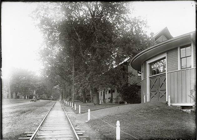 Photograph looking west at Dinkey tracks. To the right (north) the Theater Workshop (Judging Pavilion) and Landscape Architecture (Horse Barn, First) can be seen. The Farm House can be seen in the background to the left (south). Annotation: “Motor track looking west. Horse Barn (Land. Arch. Bldg.), theater pavilion on right. Farmhouse on left in background. n.d.”