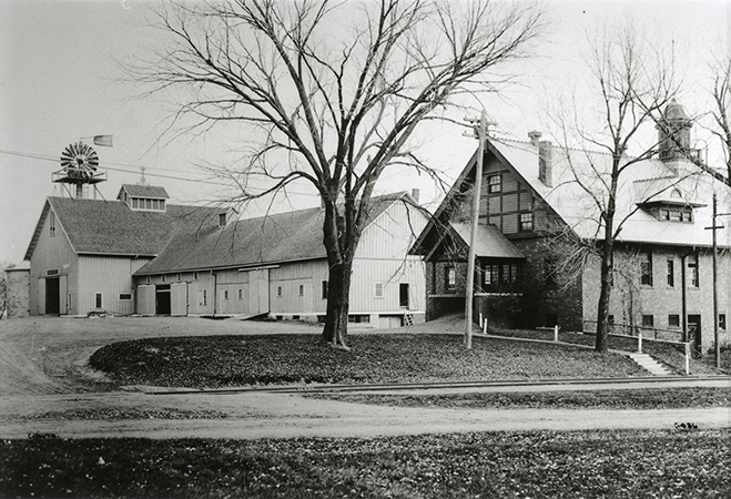 Photograph of the south and wet elevations of the Horse Barn (Landscape Architecture) on the right and the Cattle Barn in the background. The view is looking northeast. Leafless trees appear in the foreground. 