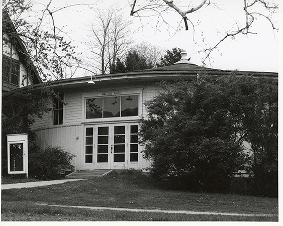 Photograph of the south door of Shattuck Theatre, facing north-northwest. There are bushes to the right of the door and a sign to the left.  