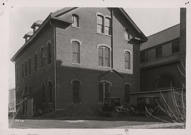 Photograph of North Hall on the Iowa State campus, showing the west and north elevations. The image depicts a three-story brick building with arched windows and adjacent structures, with early automobiles parked nearby. Annotation: North Hall 1896â€“1900 "Over Margaret Hall Kitchen."
