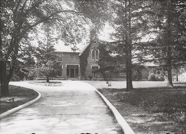 Photograph of Music Hall with surrounding landscape, showing the north elevation of the building. The image depicts a brick structure partially obscured by mature trees, with a curved driveway leading toward the entrance and landscaped grounds in the foreground. Annotation: "Old Music Hall. n.d. neg. 1310."