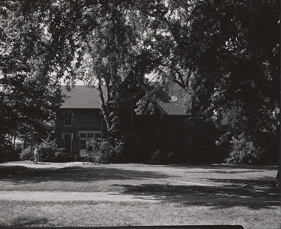 Photograph of Music Hall on the Iowa State campus, showing the north elevation. The image depicts a brick building partially obscured by mature trees, with shaded lawn in the foreground and a pathway leading toward an entrance. A person stands near the building, and a bicycle is visible nearby. Annotation: "Music Hall. 1954. neg 05120."