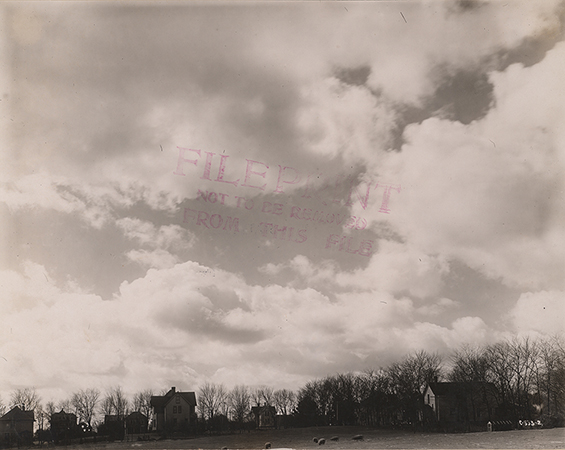 Photograph of a broad landscape view on the Iowa State campus featuring Pope Cottage among several other residential buildings. The image shows multiple houses spaced across open ground, with leafless trees in the background and a large sky filled with clouds dominating the upper portion of the photograph. Annotation: "Pope Cottage."