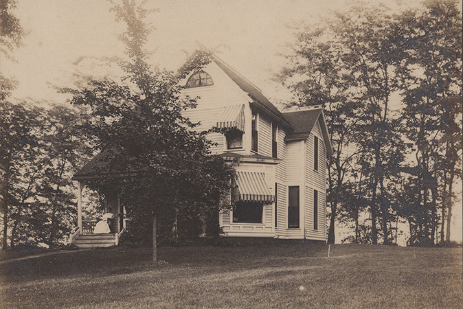 Photograph of Coburn House. It is a white two-story house with a tree in front. A woman in a white dress sits on the front porch.