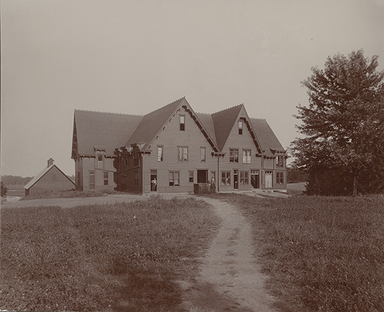 Photograph of the second creamery. It is a large three-story building. A barn sits in the backround to the left. Two men are outside the building: one leaning against a doorframe and another sitting on a box in front of a wider door. The caption reads "Creamery, 1892-1904 on site of East Hall."