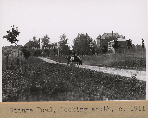Photograph of Stange Road looking to the south. A man drives a horse-drawn cart towards the south. A sign reading Iowa State College crosses the road. Teamsters Cottage #2 is visible to the left. On the right, the campanile, Agricultural Hall, and the Engineer's Cottage are visible.