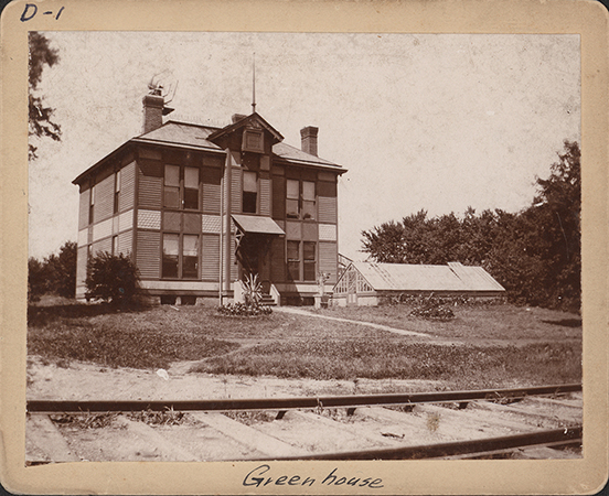 Photograph of the first agricultural experiment station and its greenhouse. It is a two story building with an awning over the front stairs. A greenhouse sits in front of it and to the right. Train tracks are in the foreground.