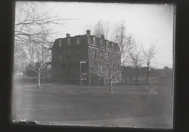 Photograph of the east boarding cottage, also known as Kirkwood Hall. The front and side of the building are shown. It is a brick building with a porch on the outside. Leafless trees surround the building.