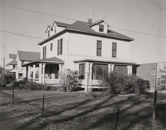 Photograph of Bevier House from the side. It has a screened in porch and a front porch. Two other houses are in the background. Part of a sign is visible in the foreground.