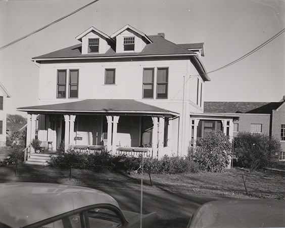 Photograph of Bevier House from the front. It is a white two-story house with a front porch. The top of two cars are in the foreground.