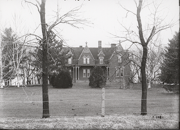 Photograph of the Gables in March, 1906. At the time it was the home of Professor Millikan Stalker. The photograph shows the front of the house with a porch. A seated swing is in the yard in front of the house. In the foreground are leafless trees and a barbed wire fence.