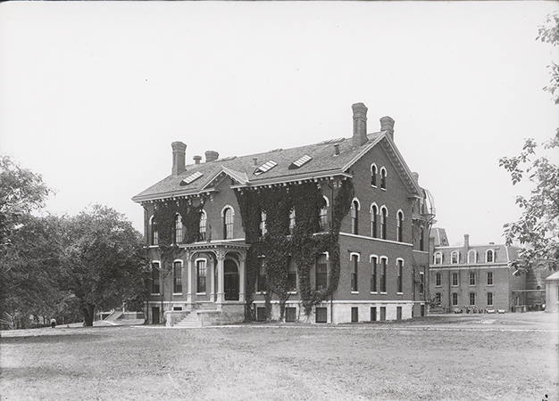 Photograph of the Chemical and Physical Laboratory. It is a two-story brick building with ivy growing on the front. The Laboratory of Mechanics is visible in the right background.