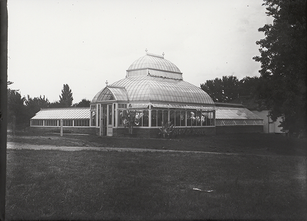 Photograph of a greenhouse on campus. It has a brick base and a barrel vaulted transept top. The caption reads, "Frame potting house where Horticulture Lab stood later from 1901-1978 on this spotl. Potting house moved to north of greenhouse."