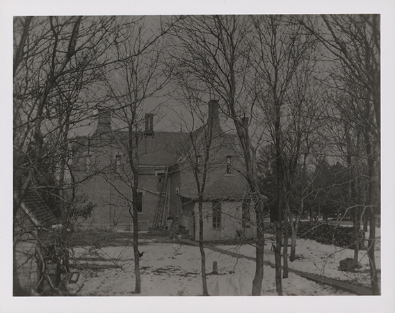 Photograph of the back of International House in winter. In the photograph, International House is in the snow, with leafless trees surrounding it. It is a brick building with an angled roof. Several windows are broken, and a ladder leans against the wall.
