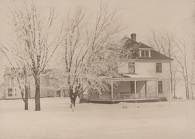Photograph of Kildee Cottage from the side. It is surrounded by snow on the ground and snowy trees. Norton House is visible in the left background.