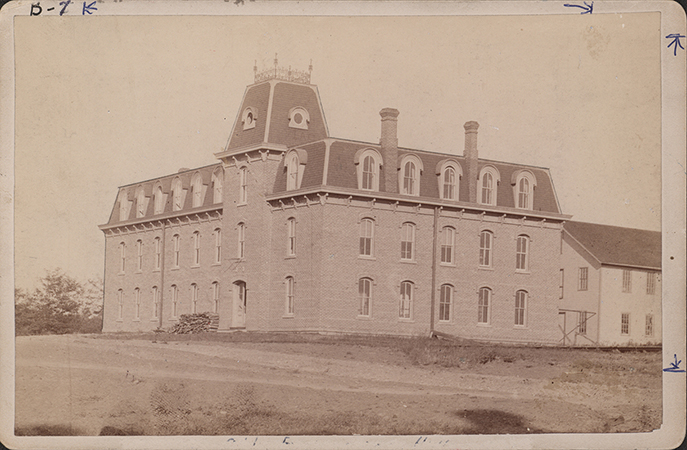 Photograph of the Laboratory of Mechanics. The photo depicts the front and side of the building. It is a brick building with three stories. A tower gable with a fence on top sits over the main entrance. A second building is partially visible to the right.