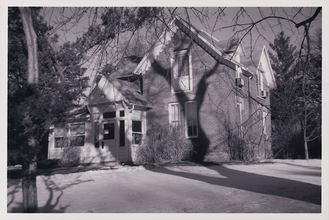 Photograph of Osborn Cottage looking north-west at the front door and east side of the building. The photograph shows trees casting shadows on the building and snow on the ground.