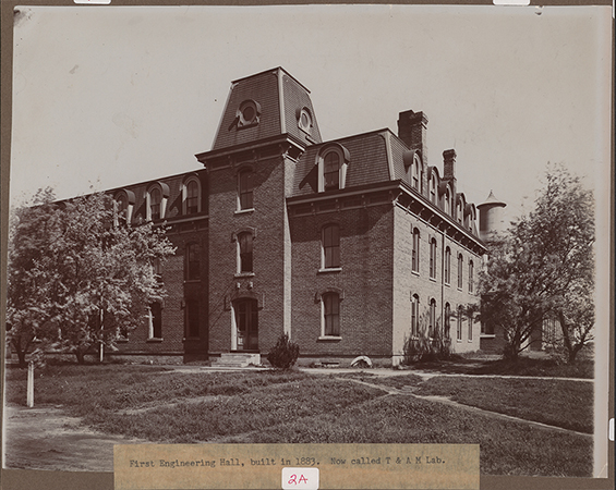Photograph looking north-west of the southeast corner of the Laboratory of Mechanics. The Marston Water Tower can be seen to the north (right). Annotation: "First Engineering Hall, built in 1883. Now called T & A M Lab."