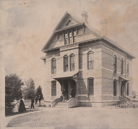 Photograph of the front and right (north) side of the English Office Building. There are eight people standing around the front and south side of the building.