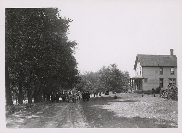 Photograph showing people and a team of horses standing in the street in front of the Engineer's Cottage. Annotation: ''Engineer's Cottage built in 1899 just east of the greenhouse. About 1915 it was moved to its present location as the eastern most cottage across from the heating plant. For some years it was the home of George Mitchell, farm superintendent.''