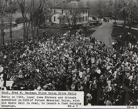 Photograph of Fred W. Beckman at the future site of the Memorial Union launching the fund raising drive for the Memorial Union. In the background cars are parked around Music Hall. Annotation: ''Prof. Fred W. Beckman stirs Union drive rally. Early in 1920, loyal Iowa Staters and friends assembled on site of future Memorial Union, with old Music Hall in rear, to launch a fund raising campaign.''