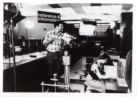 One person is using a video camera to record another person who is seated at a desk.