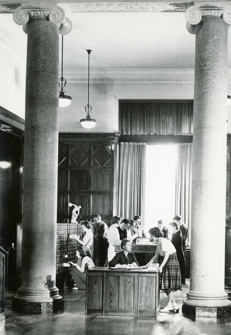 Framed by the columns in the second floor lobby, a student consults a librarian at the Card Catalog Information Desk, while many students use the card catalogs in the background.