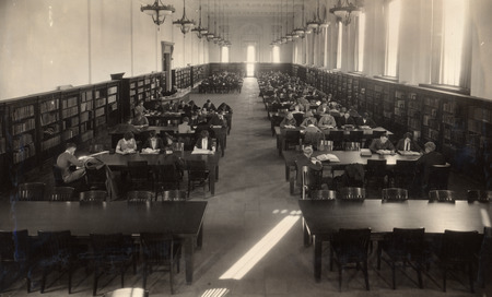 Students are studying at tables in the large Main Reading Room (currently called the Periodical Room) in the library. Two library staff members are working at the desk.