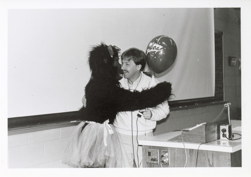 Photograph of James Noxon receiving a gift from a student in a gorilla costume on his birthday