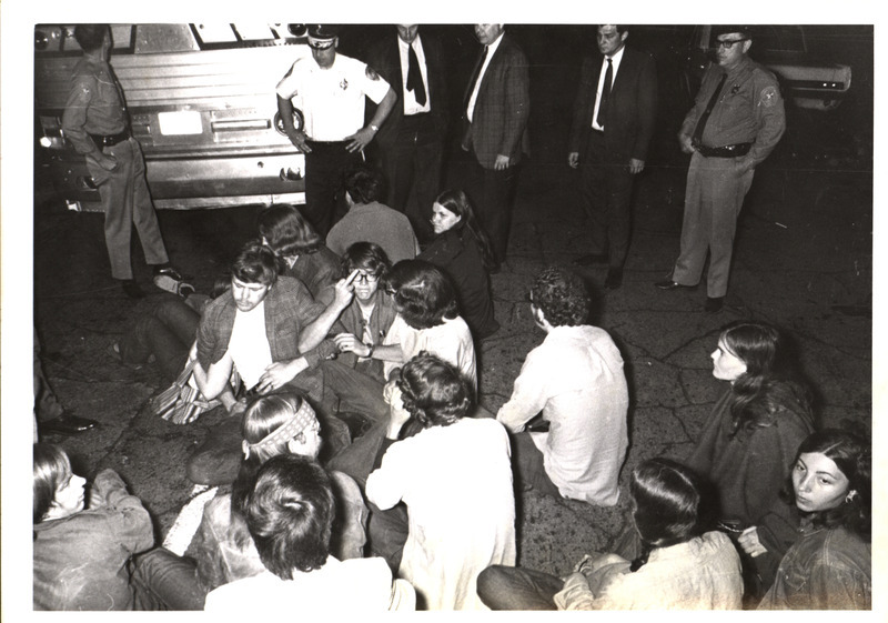 Photograph of protesters at a 1970 anti-war demonstration. Protestors are seated in the street blocking traffic, with law enforcement surrounding them.