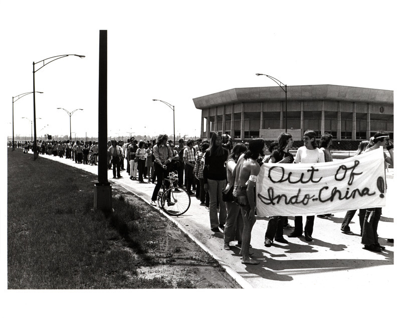 Photograph of students protesting the Vietnam War and Cambodia Invasion, lining the streets outside Hilton Coliseum. Students hold a sign reading, "Out of Indo-China!".