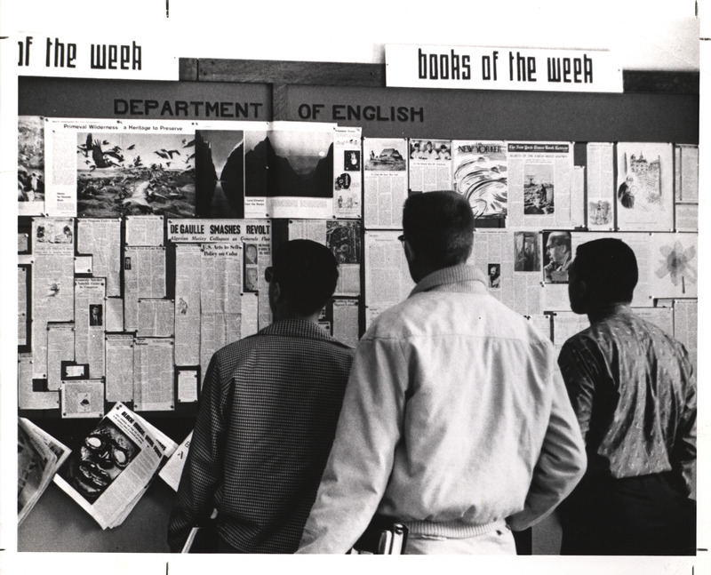 Image of students looking at the English department bulletin board, which includes clippings, literary magazines, and a sign reading "books of the week.".